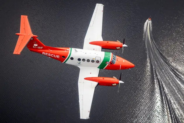 Aerial view of a twin‑engine rescue aircraft flying over open water, with a small vessel below